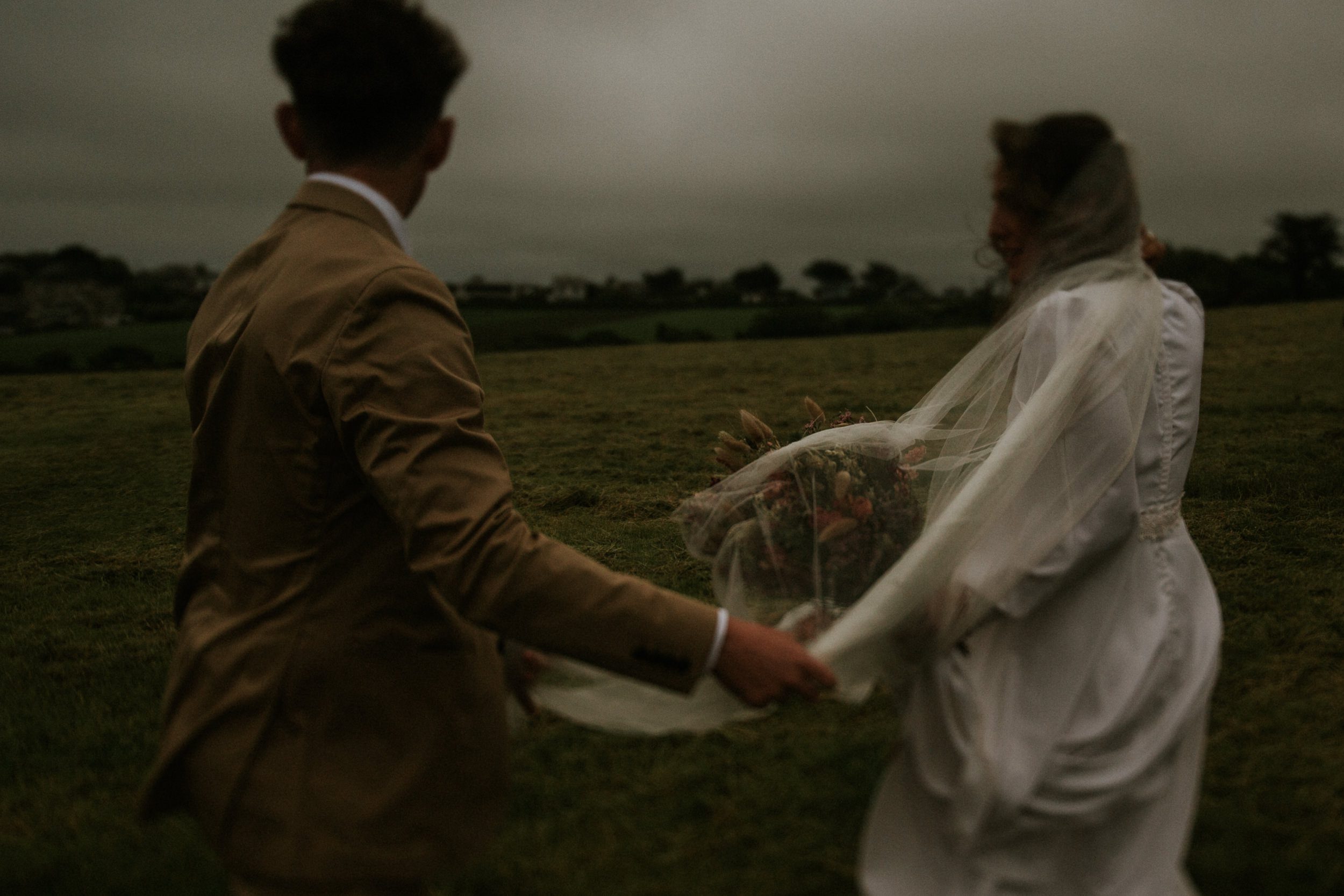 A bride and groom walk across stormy fields after their wedding at St Enodoc in Cornwall