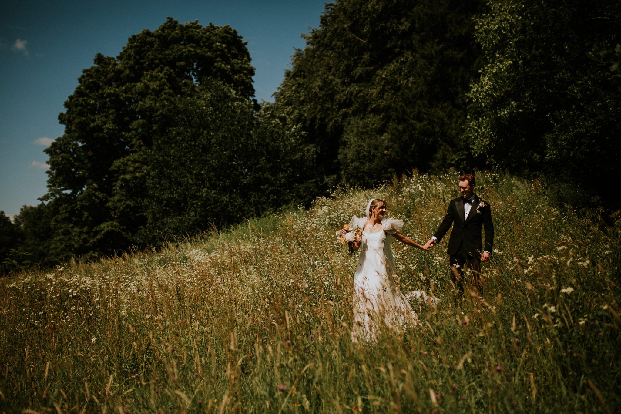 A bride and her groom chat as they walk through wildflower meadows at their wedding at The Lost Music Hall at Great Ambrook in Devon